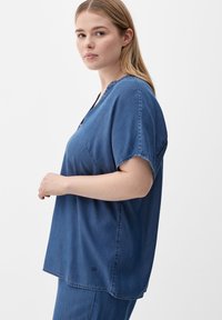 Blue denim shirt with short sleeves, a relaxed fit, and a V-neckline. Features visible stitching and a small triangle logo at the hem.