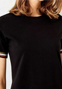 Černé tričko s krátkým rukávem, kulatým výstřihem a pruhovanými akcenty v červené, bílé a zelené barvě na lemech rukávů. Hladká textura.