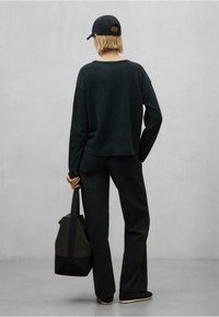 Čierna košeľa s dlhým rukávom, voľný strih, v kombinácii s čiernymi nohavicami. V jednej ruke sa drží tmavozelená a čierna taška. Na hlave má čierny klobúk.