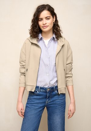 Nuori nainen kiharat hiukset, pukeutunut beigen väriseen vetoketjutakkiin, siniraitainen paita ja siniset farkut, seisoo yksinkertaisen taustan edessä.