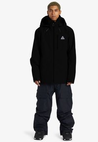 Chaqueta impermeable negra con un cuello alto y capucha ajustable; combinada con pantalones cargo oscuros con grandes bolsillos y un corte holgado.
