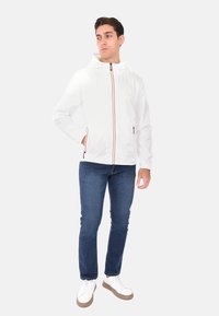 Giacca bianca con cappuccio e zip arancione, tasca frontale e polsini elasticizzati, abbinata a jeans blu e sneakers bianche.