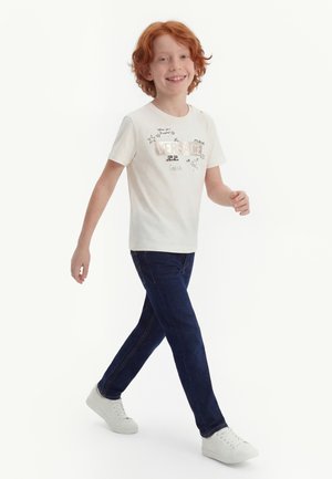 Niño pelirrojo caminando, con camiseta blanca con diseño gráfico, jeans azul oscuro y zapatillas blancas, sonriendo frente a un fondo blanco liso.