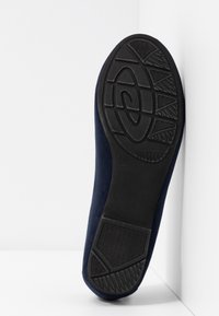 Semelle d'une chaussure plate bleu marine avec motif de grip en caoutchouc noir texturé, positionnée verticalement contre un fond blanc en coin.