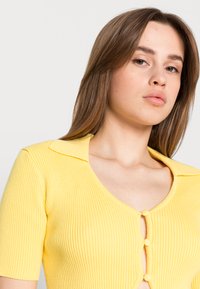 Top amarelo de malha canelada com mangas curtas, colarinho e fecho de três botões na frente, apresentando um design justo.