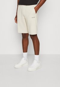 Beige Baumwollshorts mit einem elastischen Bund, seitlichen Taschen und Calvin Klein Logo. Kombiniert mit weißenSneakern und Knöchelsocken.