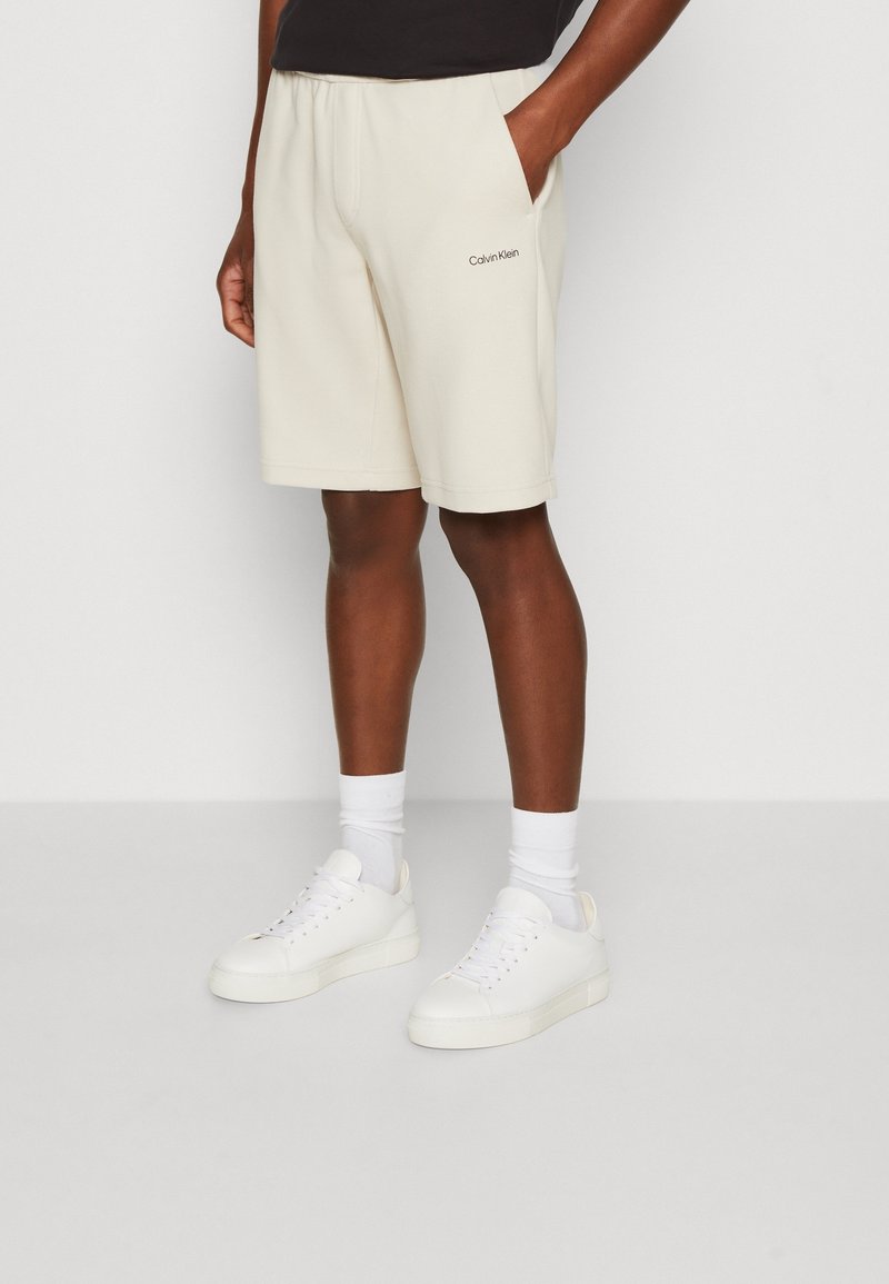 Beige Baumwollshorts mit einem elastischen Bund, seitlichen Taschen und Calvin Klein Logo. Kombiniert mit weißenSneakern und Knöchelsocken.