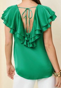 Blusa verde, con texture, con maniche a strati e balze, e una profonda scollatura sulla schiena, fissata con un nastro. Indossata con pantaloni bianchi e bracciali d'oro.