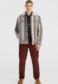 Jeune homme portant une veste rayée avec un col en laine, une chemise noire, un pantalon couleur rouille et des baskets beiges, se tenant les mains dans les poches.