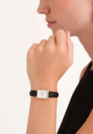Montre rectangulaire en or avec un bracelet en cuir noir. Le cadran blanc présente des index horaires et un design simple. Portée au poignet en gros plan.