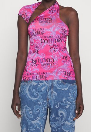 Kobieta w różowym topie bez rękawów z napisem „Versace Jeans Couture” i niebieskich ubraniach dżinsowych z kwiatowymi wzorami na białym tle.