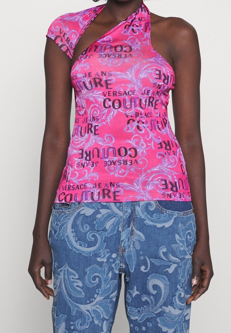 Kvinde iført ærmeløs pink top med teksten "Versace Jeans Couture" og blå mønstrede jeans med blomsterdesigns mod hvid baggrund.