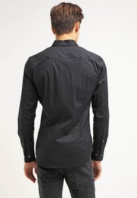 Camisa negra de botones en tejido suave, de manga larga, con un ajuste entallado, que presenta puños abotonados y un detalle de yugo en la parte posterior.