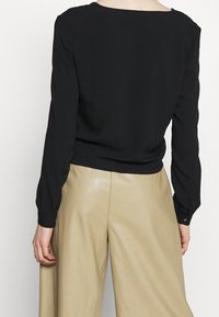 Blusa nera a maniche lunghe con scollo a barca e polsini arricciati, indossata con pantaloni beige a gamba larga. Tessuto morbido con vestibilità rilassata.
