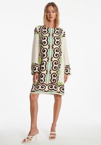 Korte mouwen, knielange jurk in crèmewit met gedurfde zwarte, groene en turquoise abstracte patronen; de stof valt zachtjes, met een losvallende pasvorm.