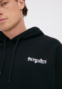 Noor mees kannab musta kapuutsiga pusa, mille rinnal on valgete ja lilla kirjaga "MEMORIZE", mida kaunistavad lillad lilled.