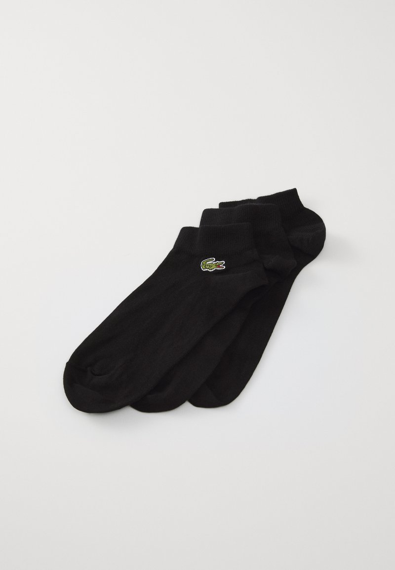 Chaussettes noires de cheville en matériau doux, avec un petit logo de crocodile brodé vert et rouge sur le côté. Quatre paires empilées.