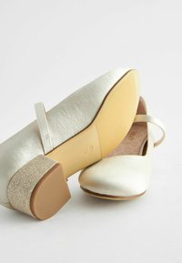 Zapatos de color dorado claro con un acabado satinado, punta redondeada y un tacón grueso cubierto de purpurina. La suela es de color marrón claro con una superficie texturizada.