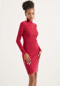 Vestito rosso aderente a maniche lunghe con colletto alto, tessuto testurizzato e spacchi laterali, accentuato da grandi orecchini ornati.