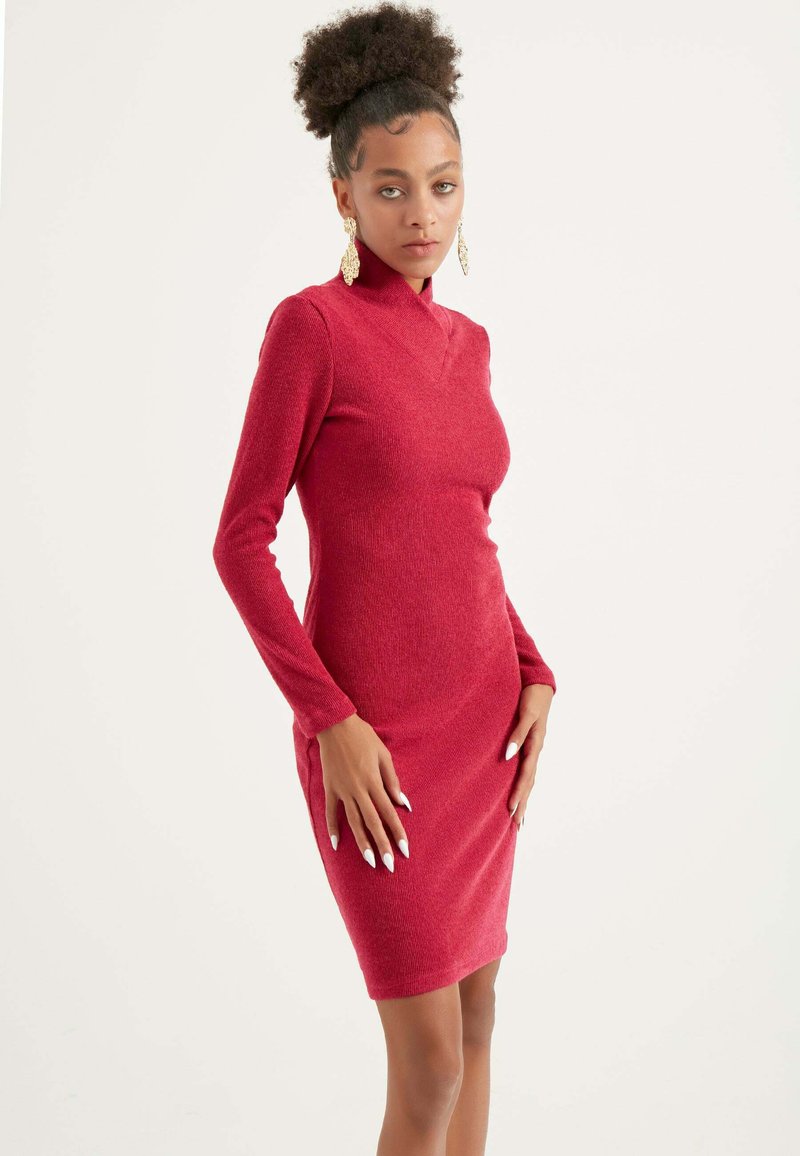 Vestito rosso aderente a maniche lunghe con colletto alto, tessuto testurizzato e spacchi laterali, accentuato da grandi orecchini ornati.