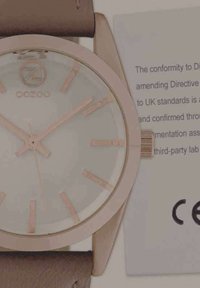 Rose gold watch with a round face, light gray dial, rose gold hour markers, brown leather strap, and a side crown for adjustment. CE mark present.