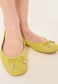 Geel leren balletschoenen met een ronde neus, voorzien van een strik en zilveren hardware-accenten, tegen een lichte achtergrond.