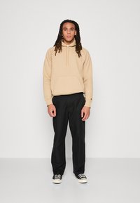 Beige hoodie met een voorzak, gecombineerd met zwarte relaxed-fit broek. De hoodie heeft een trekkoord en geribbelde manchetten. Sneakers maken de look af.