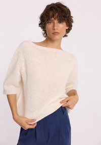 Kobieta z krótkimi kręconymi włosami, ubrana w luźny biały sweter dzianinowy i niebieskie spodnie, trzymająca hem swetra obiema rękami.