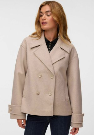 Femme portant un manteau en laine beige à double boutonnage avec de larges revers et des manches retroussées sur un haut noir et un jean foncé.