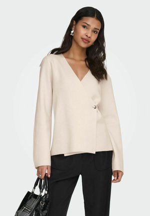 Femme portant un pull cache-cœur beige et un pantalon noir, tenant un sac à main noir texturé avec des ferrures argentées, debout devant un fond gris.