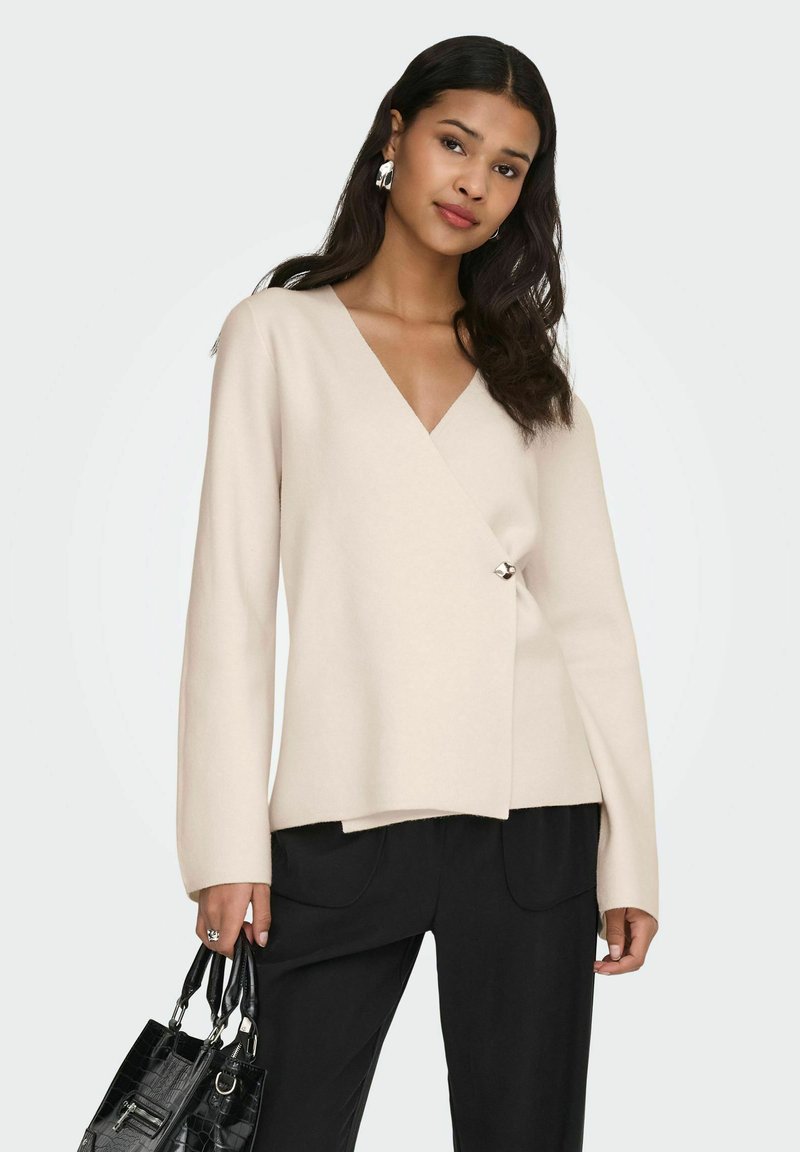 Femme portant un pull cache-cœur beige et un pantalon noir, tenant un sac à main noir texturé avec des ferrures argentées, debout devant un fond gris.