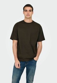 Junger Mann steht mit einer Hand in der Tasche, trägt ein schlichtes, dunkelbraunes, übergroßes T-Shirt und blaue Jeans vor einem einfarbigen Hintergrund.
