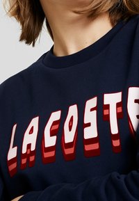 Marinblå sweatshirt med djärva, upphöjda röda och vita bokstäver av "LACOSTE". Tyget verkar slätt med rundad halsringning och långa ärmar.