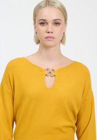 Mosterdgele trui met een ronde halslijn en een opening in de vorm van een sleuteltje. Zilverkleurige accenten aan de voorkant met een gestructureerde afwerking.