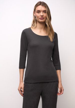 Kobieta ubrana w ciemnoszary długi rękaw z dekoltem w łódkę i pasujące szare spodnie, stojąca na jednolitym jasnym tle.