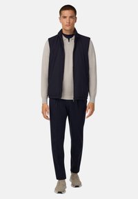 Mörkblå ärmlös väst över en ljusbeige långärmad polo, matchat med mörkblå byxor och beige sneakers. Avslappnad passform, minimal design.