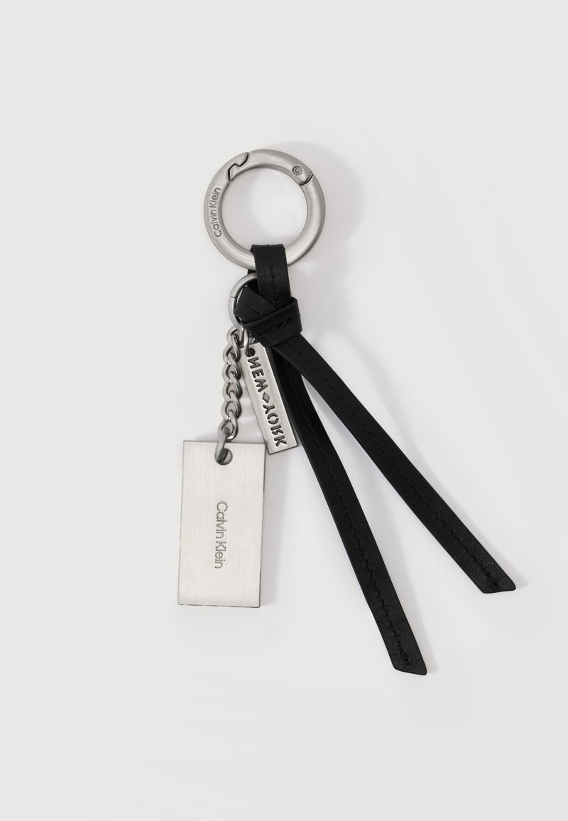 Μπρελόκ από μέταλλο Calvin Klein με ορθογώνιες πλακέτες και δύο μαύρα δερμάτινα λουριά σε λευκό φόντο.