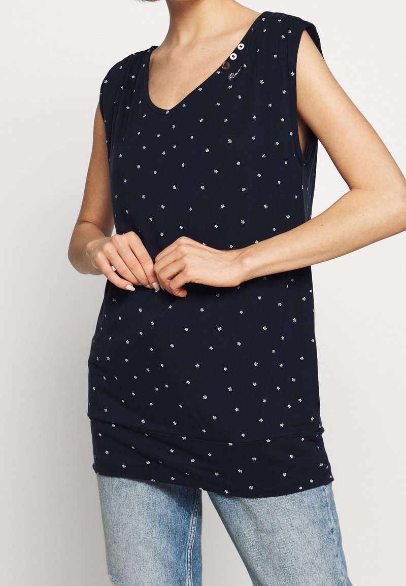 Haut sans manches bleu marine avec petit imprimé floral blanc, détails de boutons près de l'encolure, porté avec un jean bleu clair, mains délicatement jointes.