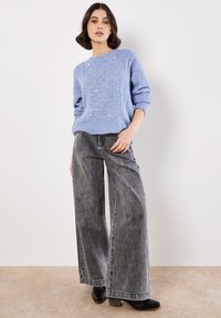 Helles blaues Strickoberteil mit Rautenmuster, kombiniert mit hochgeschnittenen, weit geschnittenen grauen Jeans. Schwarze, Absatz-Ankle-Boots.