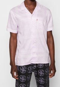 Man wearing a light purple short-sleeve button-up shirt with subtle paisley print and black shorts with circular patterned text.