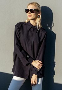 Black button-up shirt with long sleeves, featuring starfish-shaped cuff accents. Worn with white pants against a neutral background.