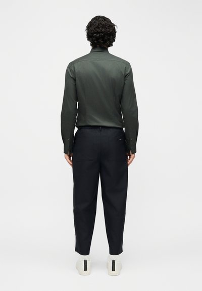 Άνδρας με σγουρά μαλλιά, φοράει μακρυμάνικο σκούρο πράσινο πουκάμισο, μαύρο παντελόνι με πίσω τσέπες και λευκά αθλητικά παπούτσια με μαύρες ρίγες.