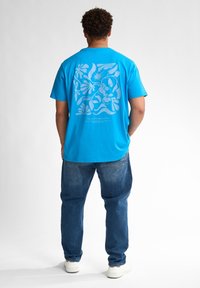 Modrá bavlněná tričko s velkým bílým květinovým motivem na zádech, spárované s volnými modrými denimovými džíny a bílými teniskami.