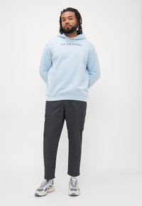 Light blue sweatshirt with hood and black text "JACK & JONES," paired with dark cargo pants and gray sneakers, standing against a white background.