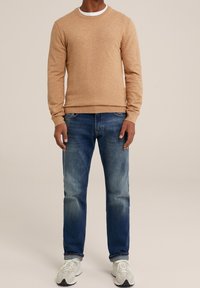 Ljusbrun stickad tröja med rund hals, matchad med blå denimjeans som har lätt blekna områden. Vita sneakers fulländar outfiten.