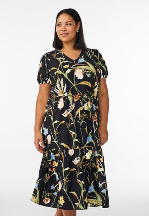 Frau trägt ein schwarzes Midikleid mit Blumenmuster, Puffärmeln und V-Ausschnitt, steht vor einem schlichten weißen Hintergrund.