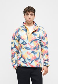 Νεαρός άνδρας που φοράει πολύχρωμο φλις πουλόβερ με αφηρημένα σχέδια και μερικώς κουμπωμένο γιακά, στέκεται μπροστά σε απλό λευκό φόντο.