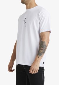 Camiseta blanca de algodón con mangas cortas, escote redondo y un pequeño estampado gráfico negro en el pecho; combinada con pantalones negros.