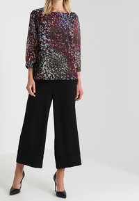 Haut noir transparent à motif floral multicolore, associé à un pantalon noir à jambes larges et des talons noirs.