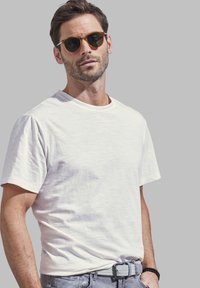 Weißes T-Shirt mit Rundhalsausschnitt aus leichtem Stoff, kombiniert mit grauen Jeans und einem strukturierten Gürtel, ergänzt durch runde Sonnenbrillen.